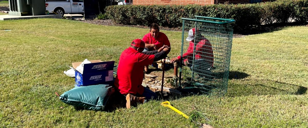 3 men in red shirts are performing modifications to an existing lawn sprinkler system